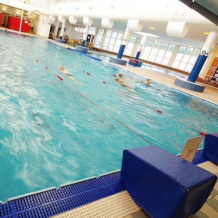 Indoor swimming pool at a leisure centre with swimmers in action, separated by lane ropes, viewed from a poolside angle with blue mats on the deck.