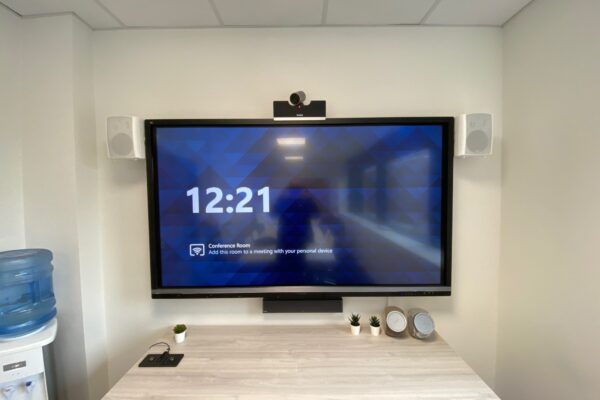 A modern conference room setup displaying a large screen showing the time "12:21" and a message about connecting to a meeting, flanked by two speakers, in a clean and organized space with advanced audio visual systems.