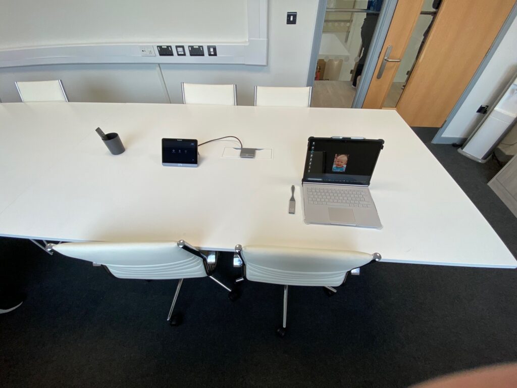 A modern conference room with a large white table, four chairs, and a laptop on one end next to a small audio visual system speaker device.