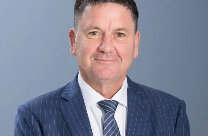 Portrait of a middle-aged man in a blue pinstripe suit and ICO tie smiling against a light blue background.