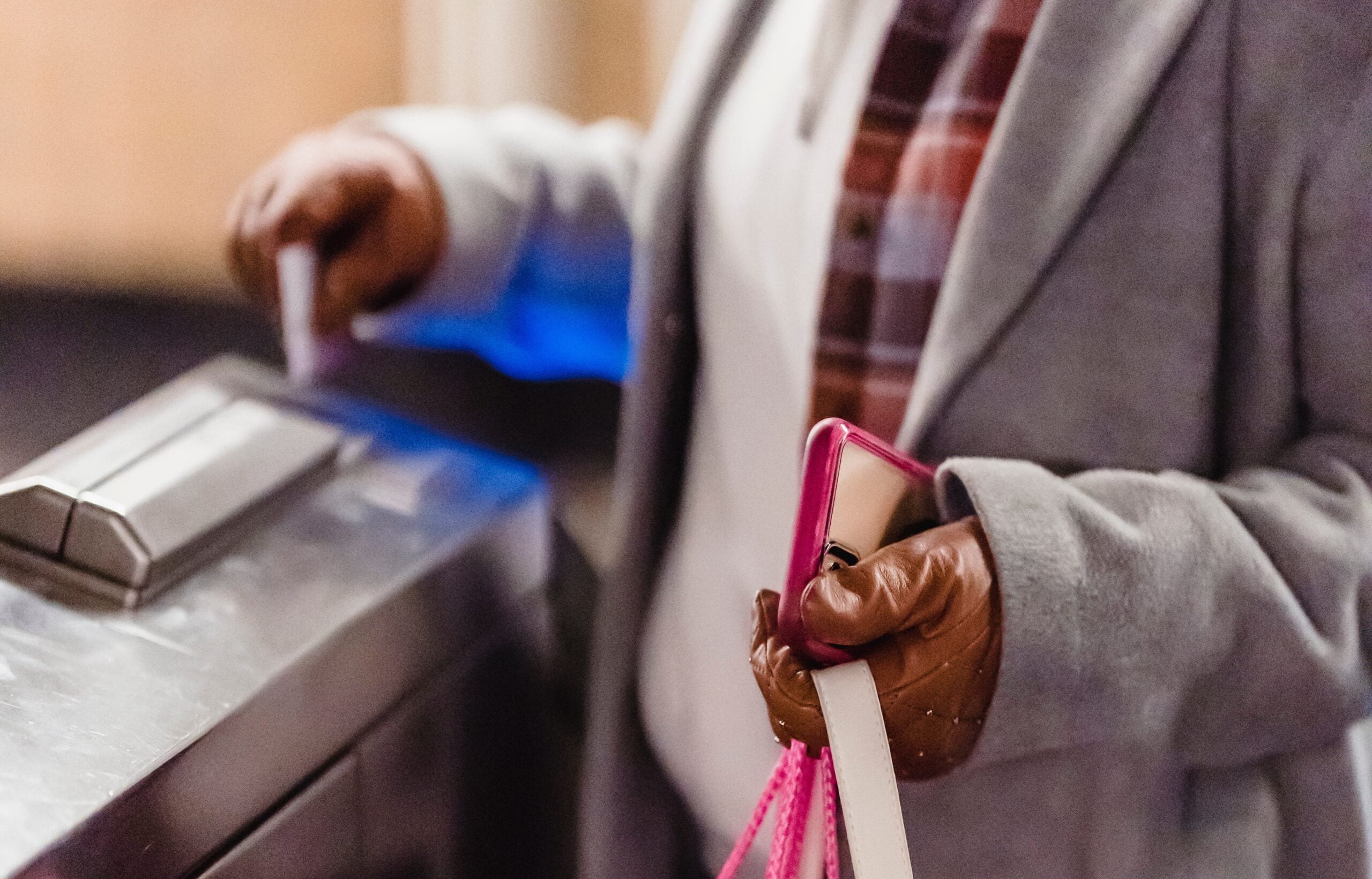 A person wearing a plaid coat and brown gloves uses a credit card on a business access control system while holding a smartphone and shopping bags.