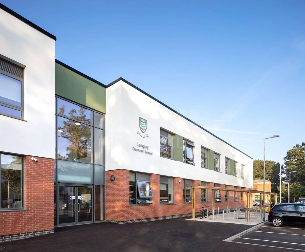 Modern two-story educational building with large windows, brick facade on lower level, and parked cars on a sunny day.