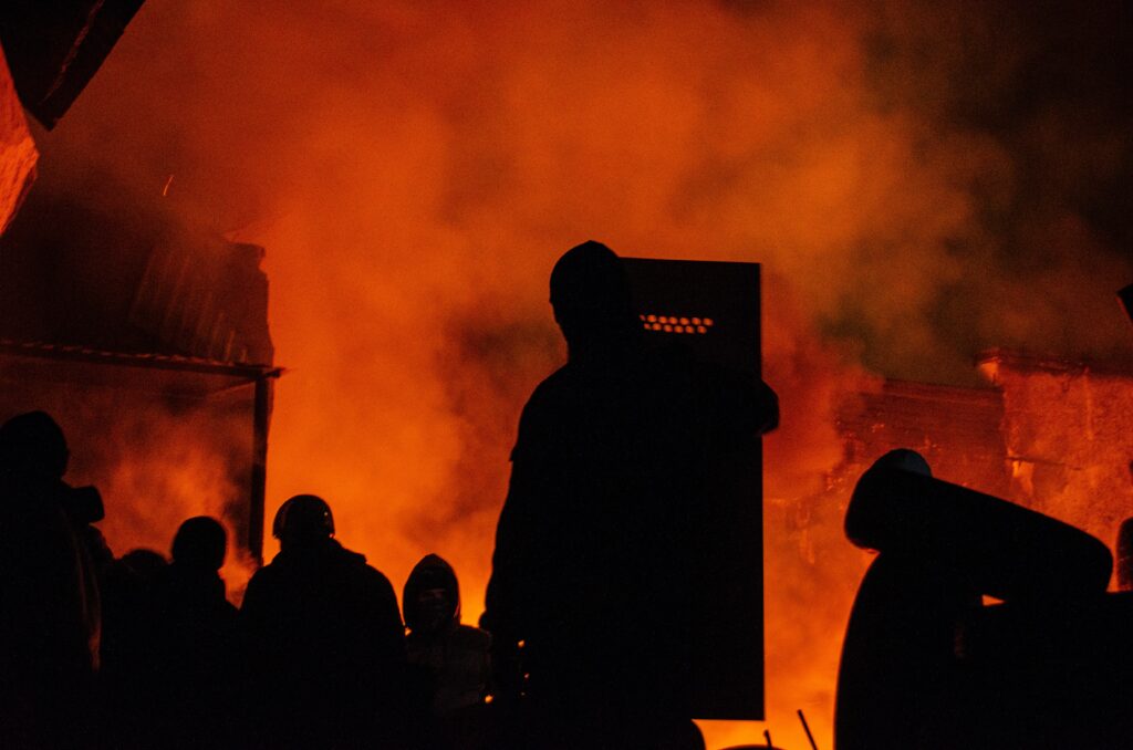 Silhouetted figures standing in front of a large fire at night, with orange flames and smoke symbolizing IT disaster recovery services filling the air around them.