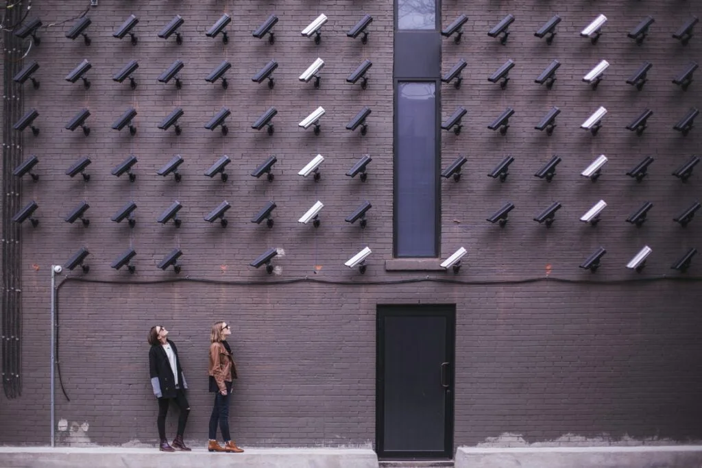 Two people walking past a dark building wall covered with numerous cctv cameras.