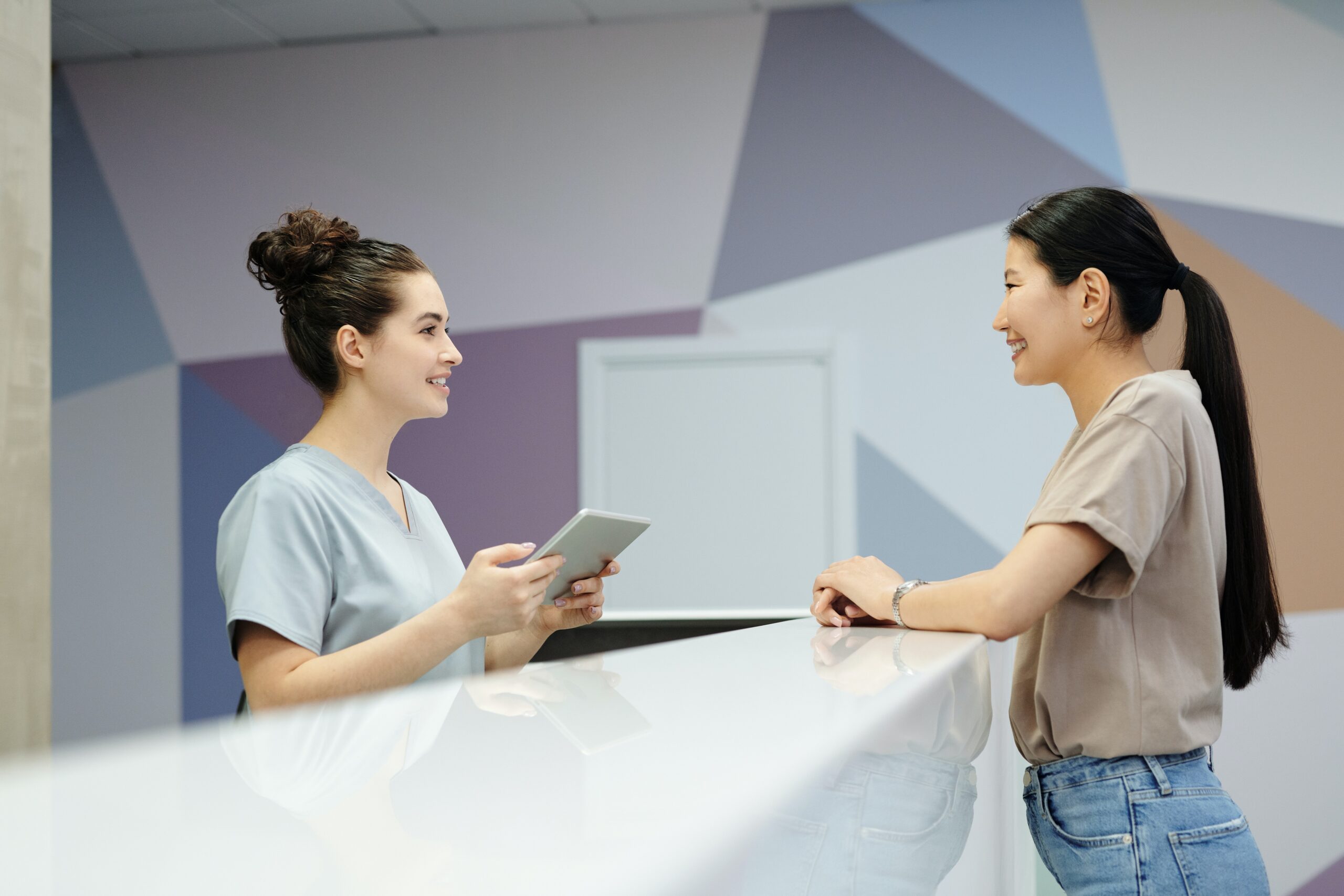 Two women discussing business access control systems at a reception desk, one holding a tablet and the other standing, in a room with a geometric wall design.