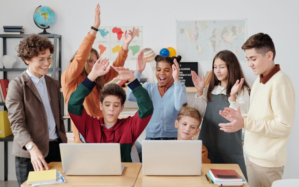 Teacher and students celebrating with raised hands in a classroom equipped with laptops and a world map on the wall, benefiting from their school's backup solution.
