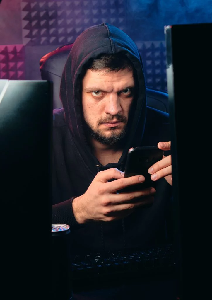 A man in a hooded jacket looks intently at his smartphone, possibly researching watering hole attacks, surrounded by two computer monitors in a dimly lit room with a blue backdrop.