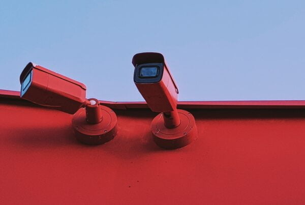 Two cctv cameras mounted on a red wall under a blue sky.