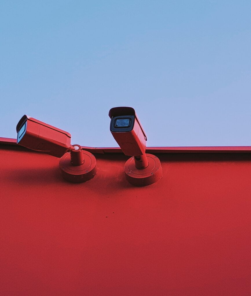 Two cctv cameras mounted on a red wall under a blue sky.