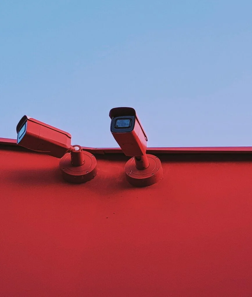 Two cctv cameras mounted on a red wall under a blue sky.