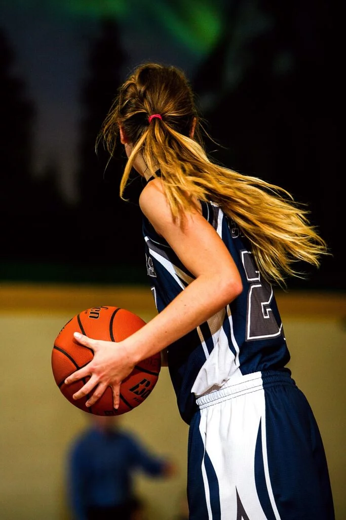 A female basketball player in a dark uniform dribbles the ball down the court, her ponytail flying behind her, with a video wall displaying the game in the background.