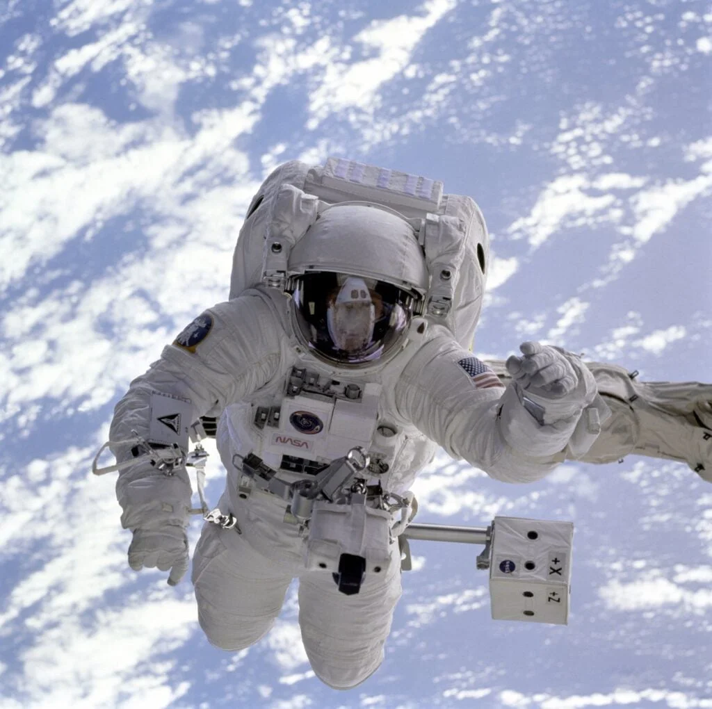An astronaut in a white nasa spacesuit conducting a spacewalk, with earth and clouds visible in the background.