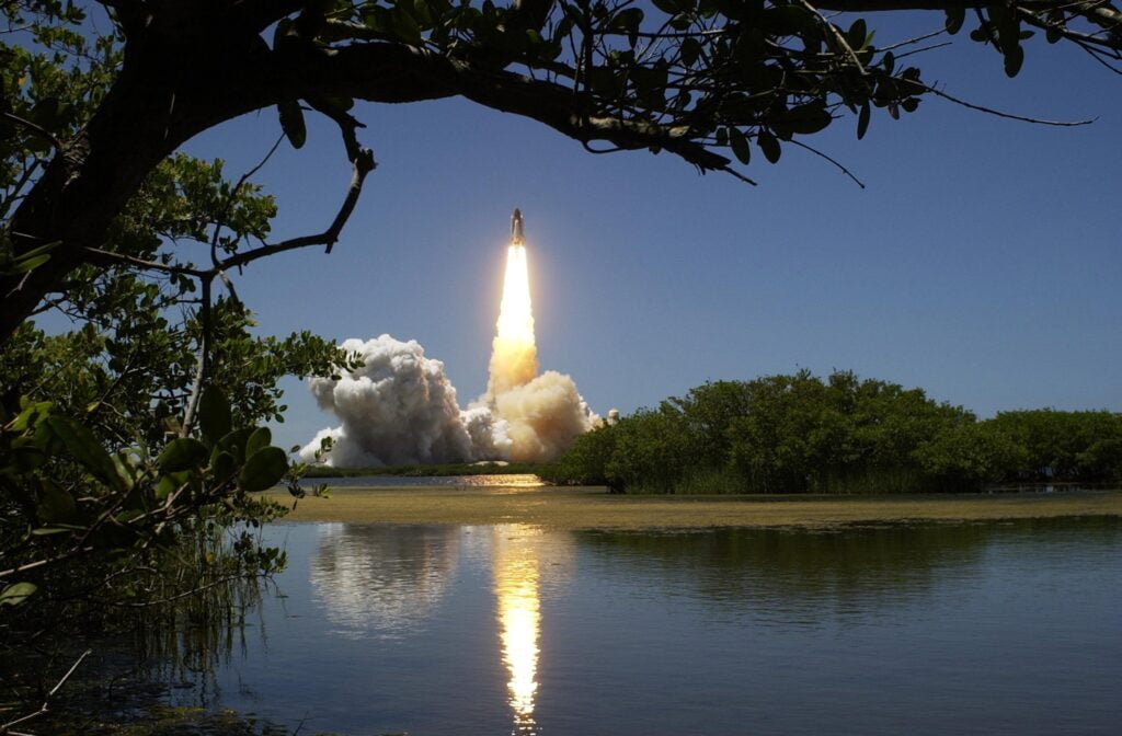 Space shuttle launching over a calm lake, viewed from behind lush green foliage under a clear sky.