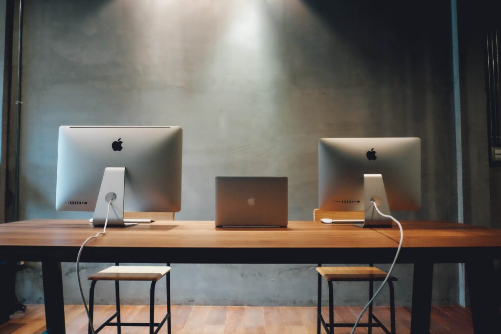 Two apple imacs and a macbook procured through IT procurement services, on a wooden table in a dimly lit room with concrete background.
