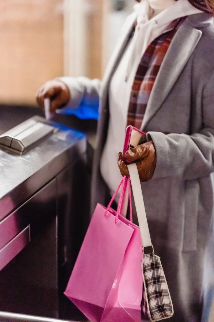 Woman in a coat and gloves using a credit card at a business access control system, holding a pink shopping bag and a plaid wallet.