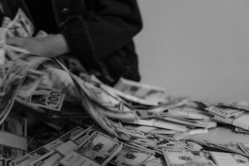 Black and white image of a person's hand shuffling through a large pile of 100 dollar bills.