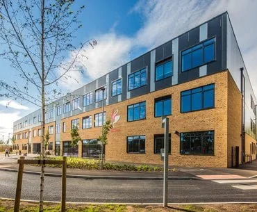 Modern three-story building with a combination of yellow brick and gray paneling under a blue sky, surrounded by young trees and landscaping.