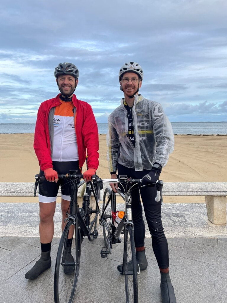 Two male cyclists, wearing helmets and sportswear, standing with their bicycles on a beachfront promenade under an overcast sky.