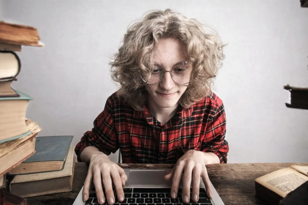 A young person with curly hair wearing glasses and a red plaid shirt typing on a laptop, surrounded by stacks of books on a wooden table.