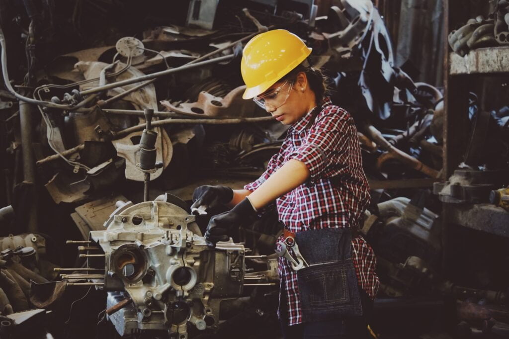 A woman mechanic in a hard hat and glasses works on a car engine in a cluttered workshop, utilizing the Help To Grow initiative to enhance her skills.