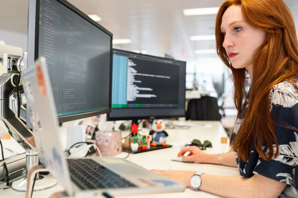 A red-haired woman works intently at her office desk with multiple monitors displaying code, as part of the Help To Grow initiative.