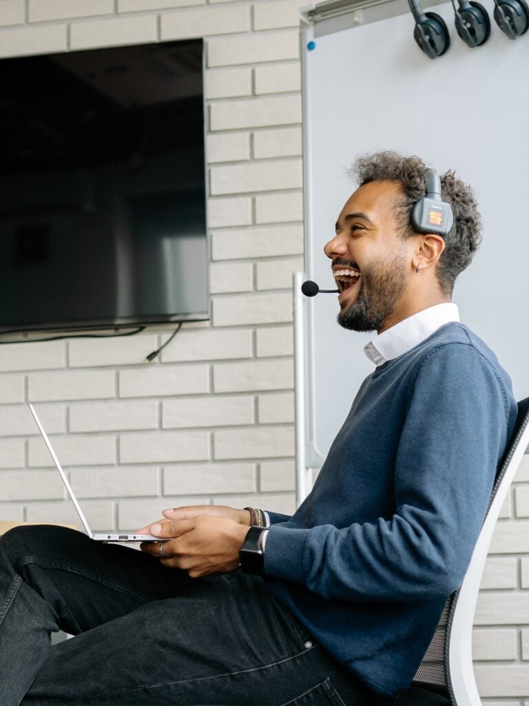 A man wearing a headset laughs while sitting on a chair with a laptop on his lap, in an office setting with a whiteboard and tv in the background, likely participating in an Infrastructure Technician Apprenticeship.
