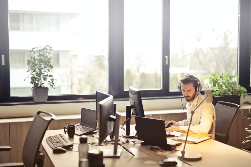 A man wearing headphones works intently at a computer in a bright, modern office with large windows and unified communications systems.