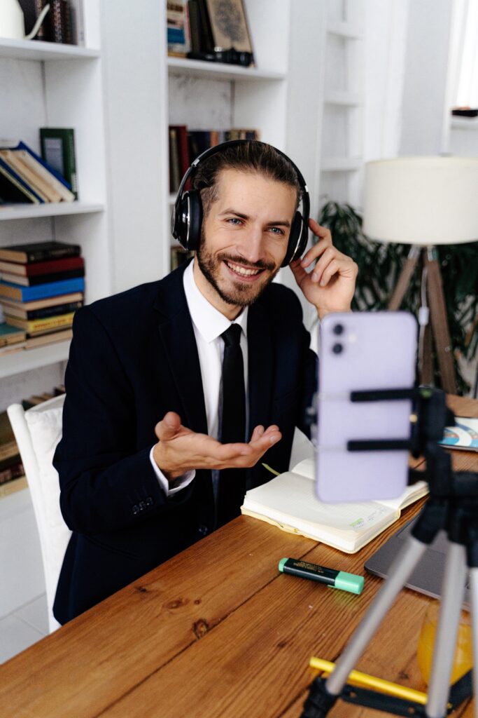 A man in a suit wearing headphones uses a VoIP phone on a tripod to conduct a video call from a desk filled with books and papers.