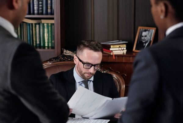 A focused man in glasses reviews IT procurement documents at a conference table with two standing colleagues in a professional, wood-paneled office.