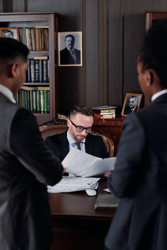 A focused man in glasses reviews IT procurement documents at a conference table with two standing colleagues in a professional, wood-paneled office.