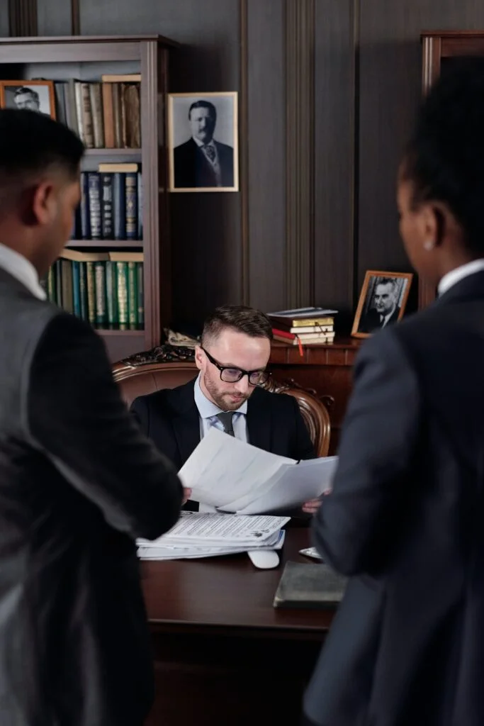 A focused man in glasses reviews IT procurement documents at a conference table with two standing colleagues in a professional, wood-paneled office.