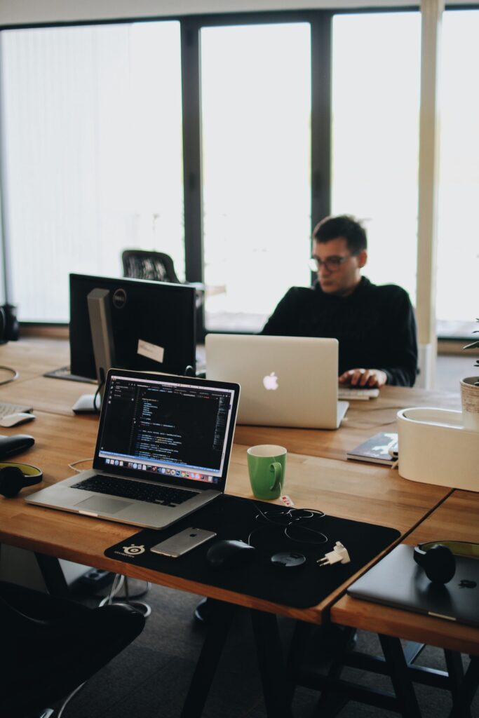 Man working on a laptop in a modern IT services office with multiple computers and a large window in the background.