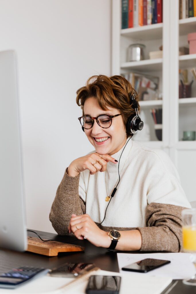 A woman wearing headphones smiles while looking at her computer screen in a home office with 3CX phone systems and books in the background.