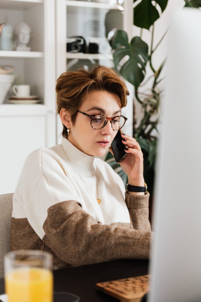 A woman in glasses talks on a 3CX phone system while looking at a computer screen, seated at a kitchen table with a drink beside her.