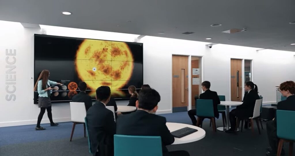 A classroom with students watching a teacher present an image of the sun on a large screen labeled "science.
