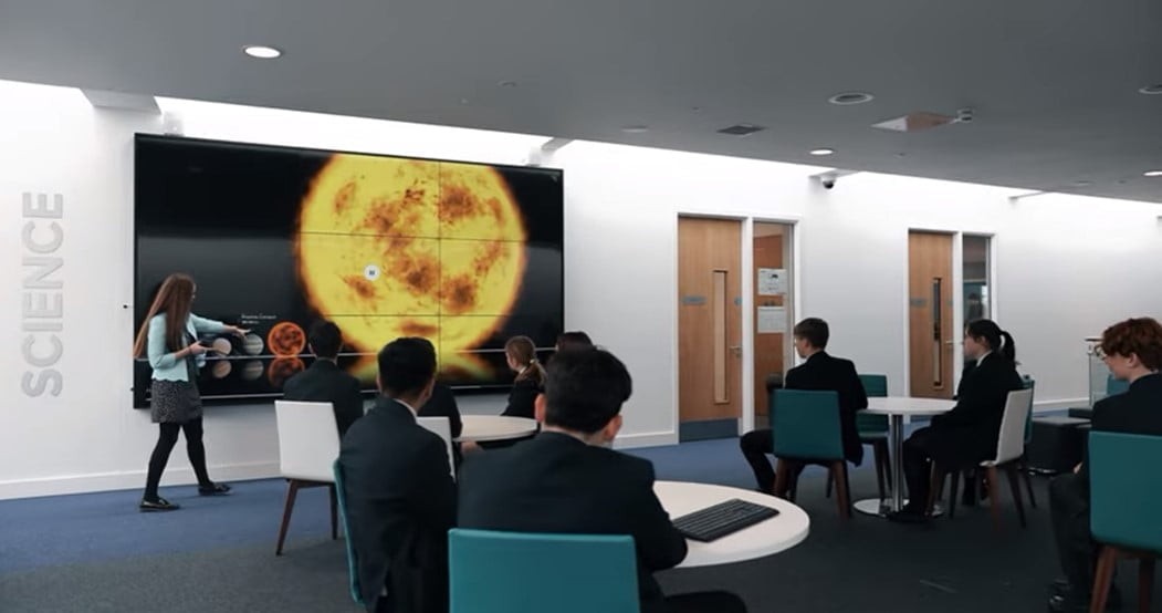 A teacher presenting a solar system lesson on a large screen to students seated at desks in a modern classroom.