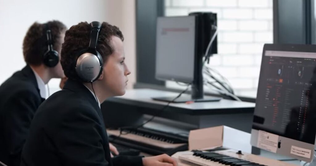 A man in headphones intently uses a digital audio workstation with a midi keyboard at a desk in a modern office setting.