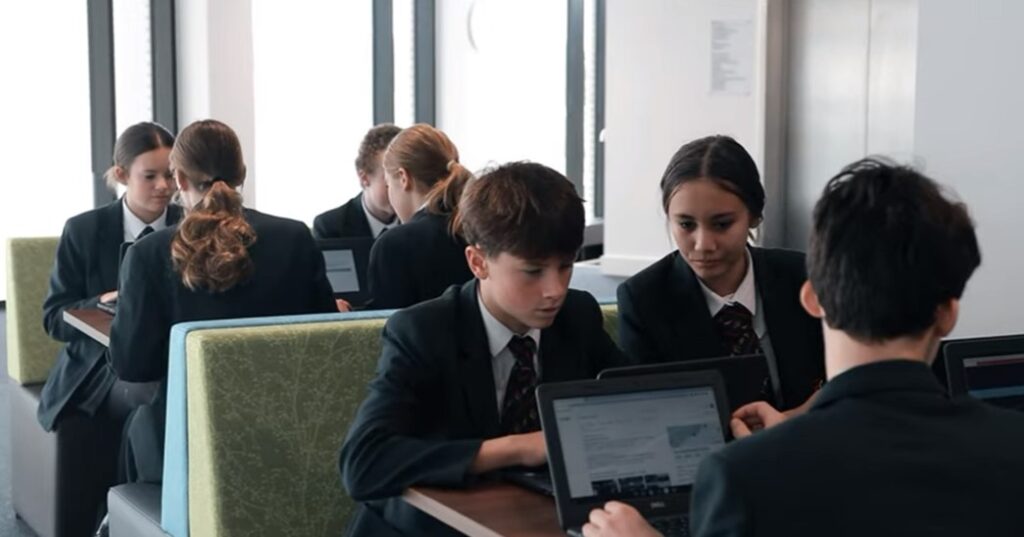 Students in school uniforms working on laptops at a table in a classroom, discussing and concentrating on their screens.