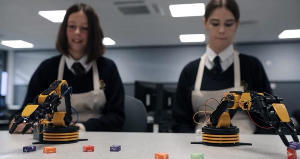 Two students in uniforms experimenting with robotic arms sorting colorful cubes on a desk in a classroom.