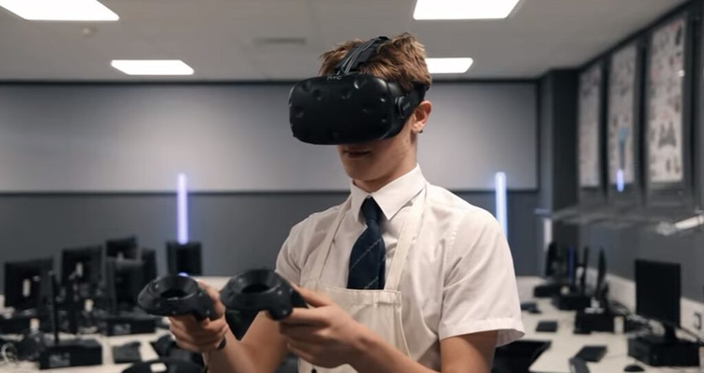 A young man in a shirt and tie uses a vr headset and controllers in a classroom with computers.