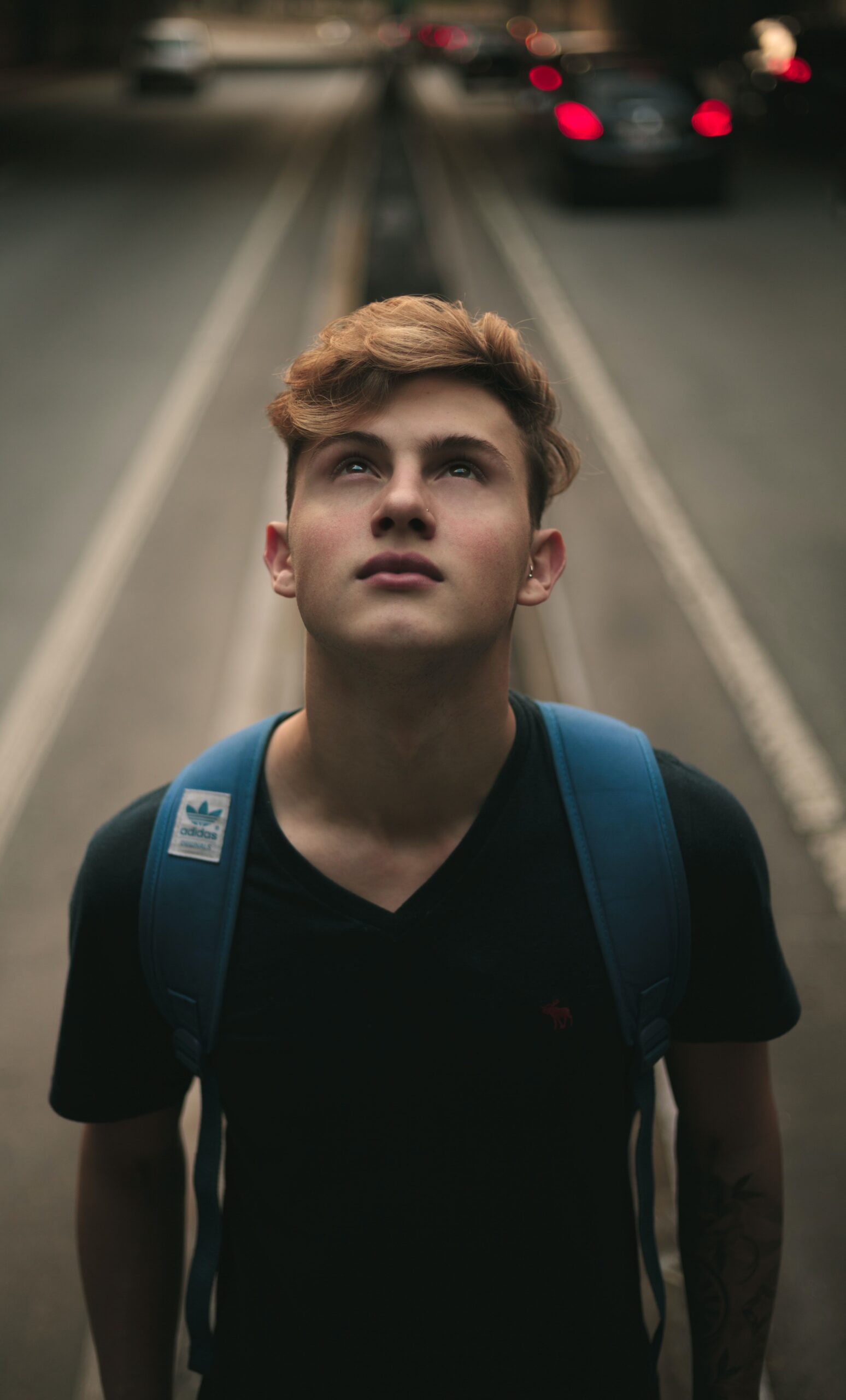 Young man with a backpack standing on a street, looking upwards with a thoughtful expression, cars and tram tracks blurred in the background.