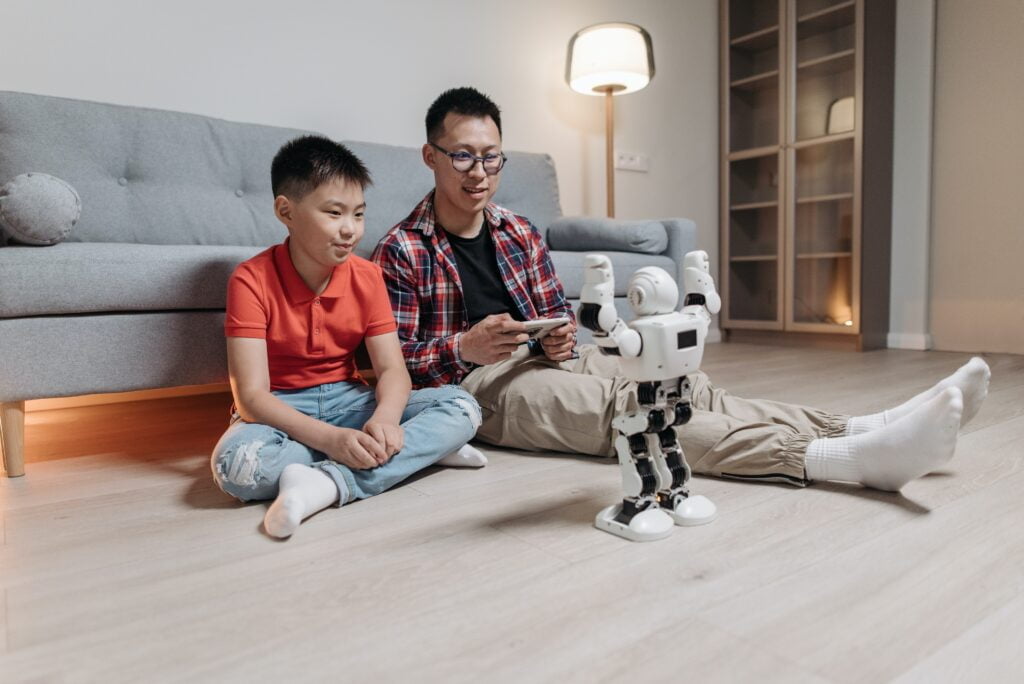 Father and son sitting on a living room floor, interacting with a humanoid robot next to a plush gray sofa.