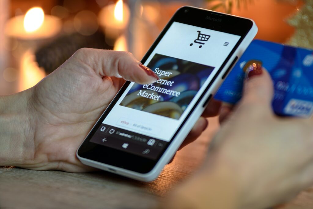 Person shopping online using a smartphone with a credit card in hand, displaying an e-commerce website on their business broadband connection. Warm, blurred background lights enhance the festive ambiance.