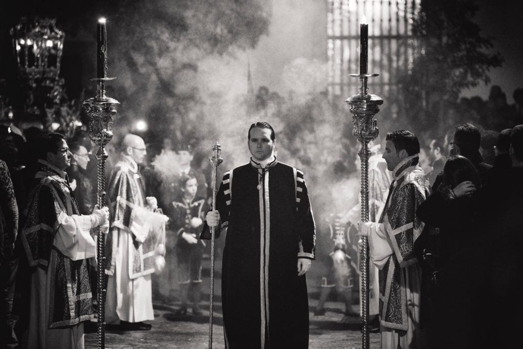 A solemn religious procession at night with people holding candlesticks and an incense bearer, in black and white.