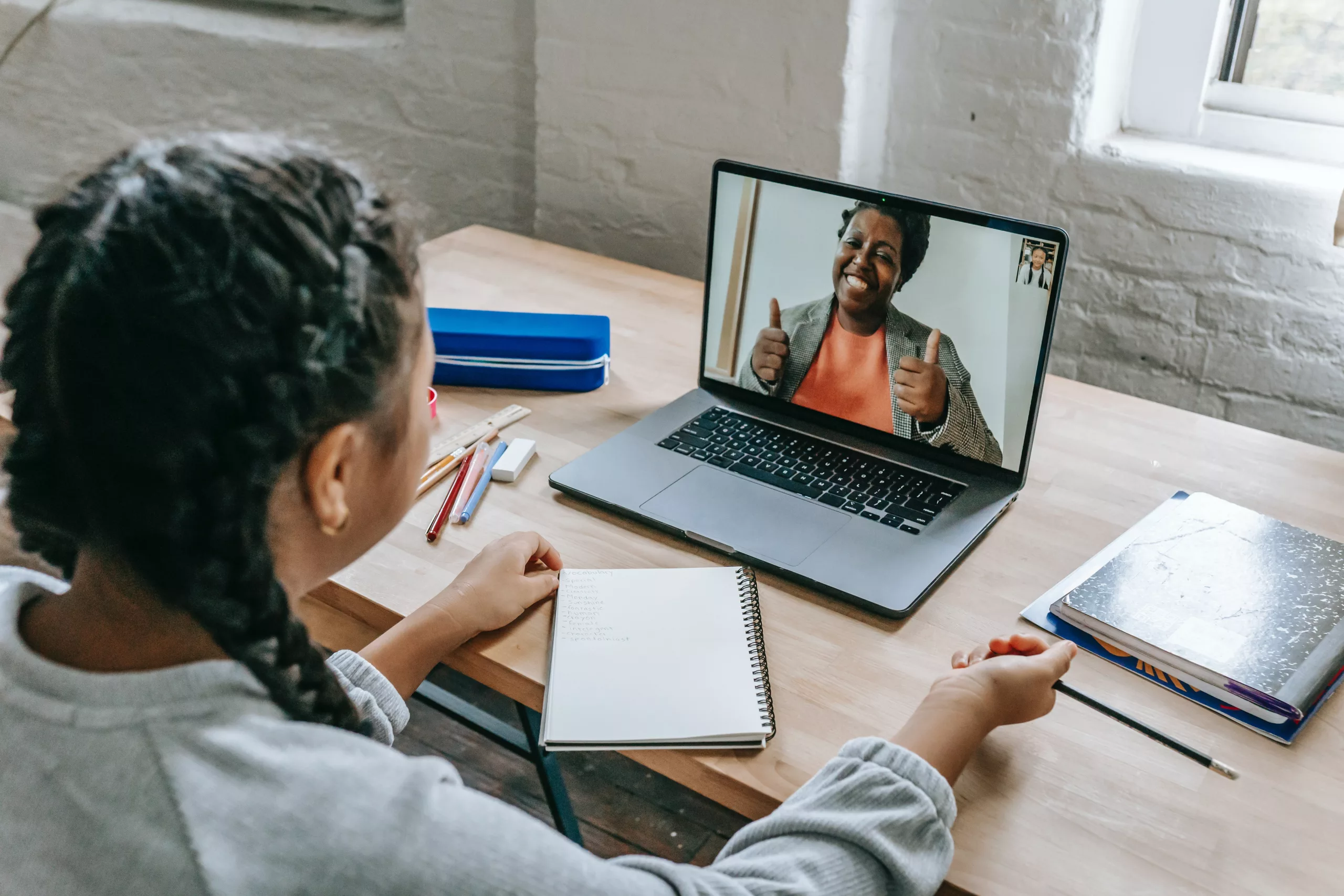 A young girl participates in an online class, attentively watching a happy teacher giving a thumbs up on her laptop screen.