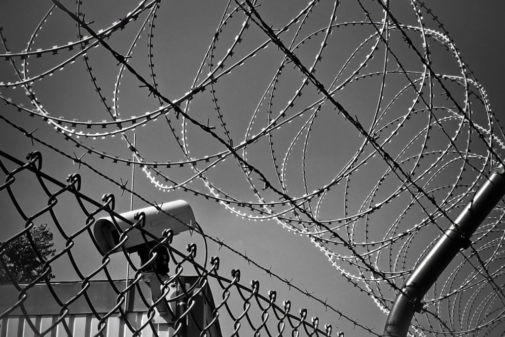 Black and white image of barbed wire coiled along the top of a chain-link fence against a sky backdrop.