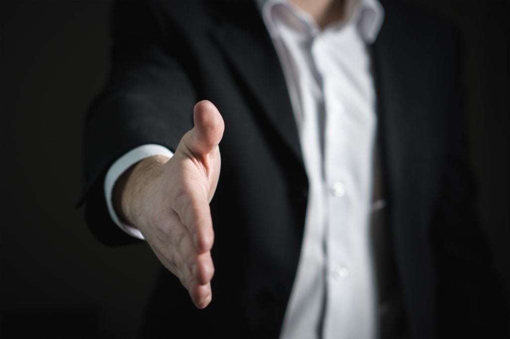 A man in a dark suit with a cyber essentials certification badge extending his hand for a handshake, focus on the hand with a blurred background.