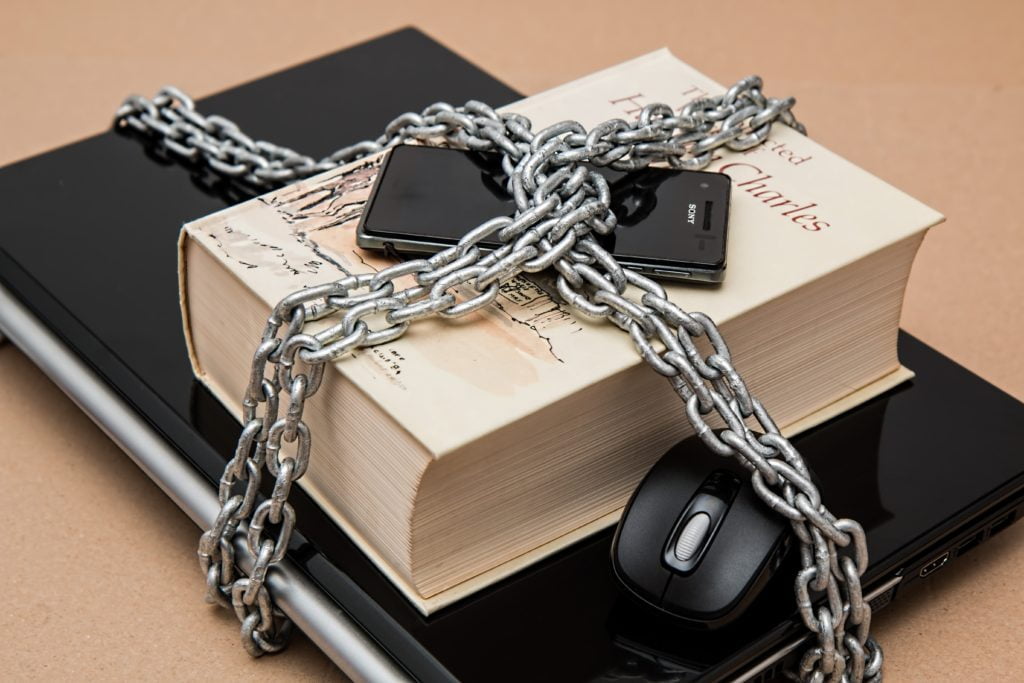 A book titled "selected plays of Charles," a smartphone, and a computer mouse secured with a metal chain on a tan background to secure SaaS apps.