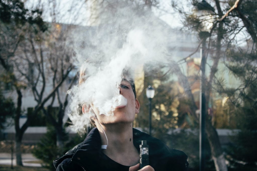 Young man exhaling vapor from an electronic cigarette outdoors, surrounded by trees.