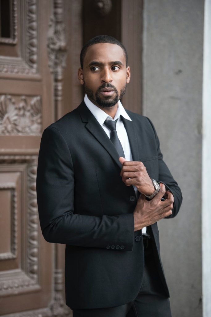 A man in a black suit and tie adjusting his cufflink while standing by a carved wooden door, possibly before meeting with clients about penetration testing services.